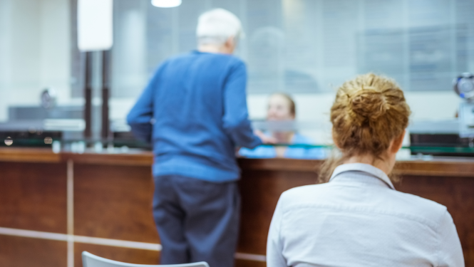 Klients pie kases vai klientu apkalpošanas lodziņa bankā, pastā vai valsts iestādē, fonā redzama darbiniece, bet priekšplānā – cita kliente no mugurpuses.
