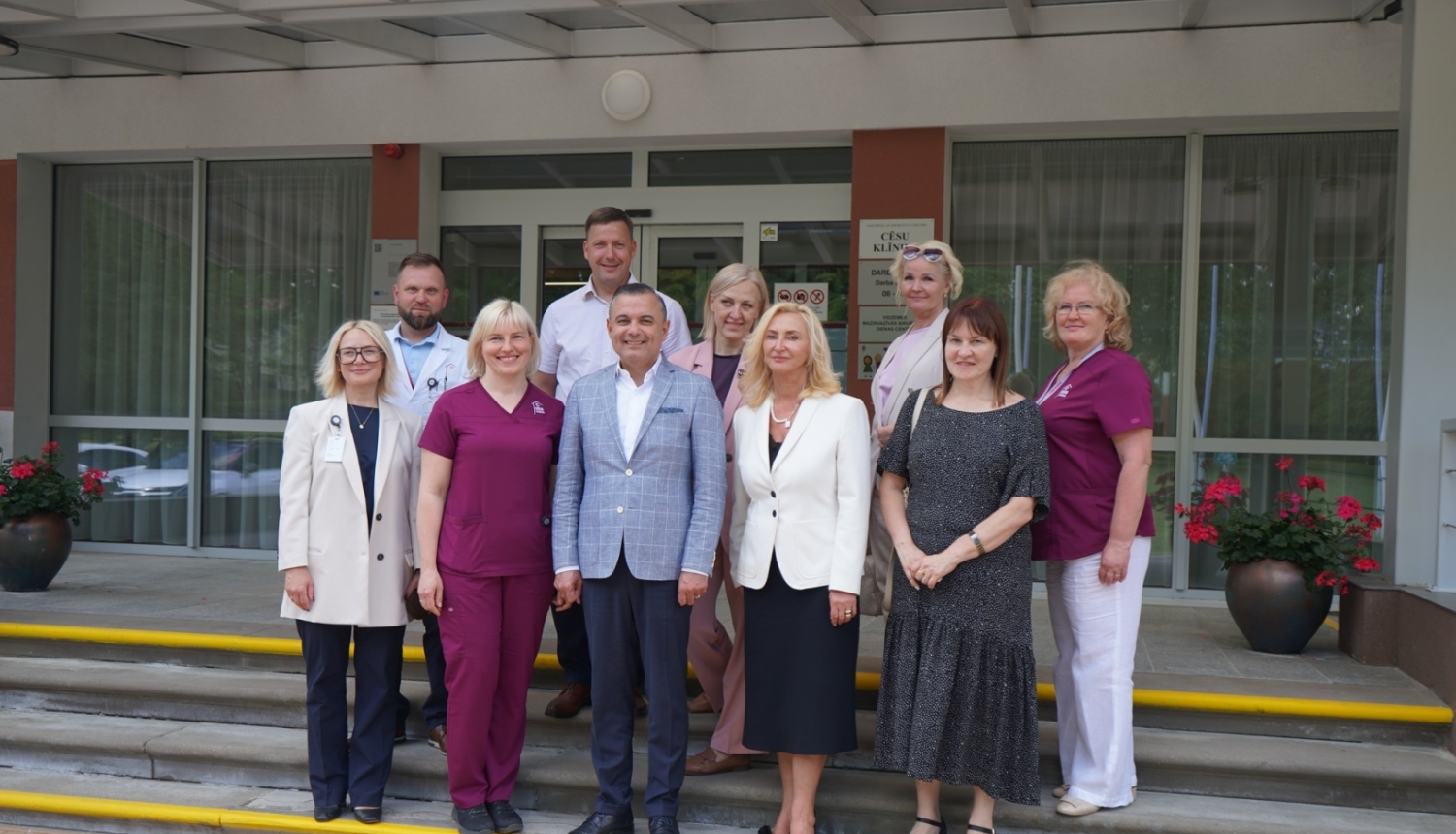 Grupas fotoattēls no reģionālās vizītes pie Cēsu klīnikas ieejas, kurā redzami klīnikas pārstāvji un viesi kopīgā foto uz kāpnēm.