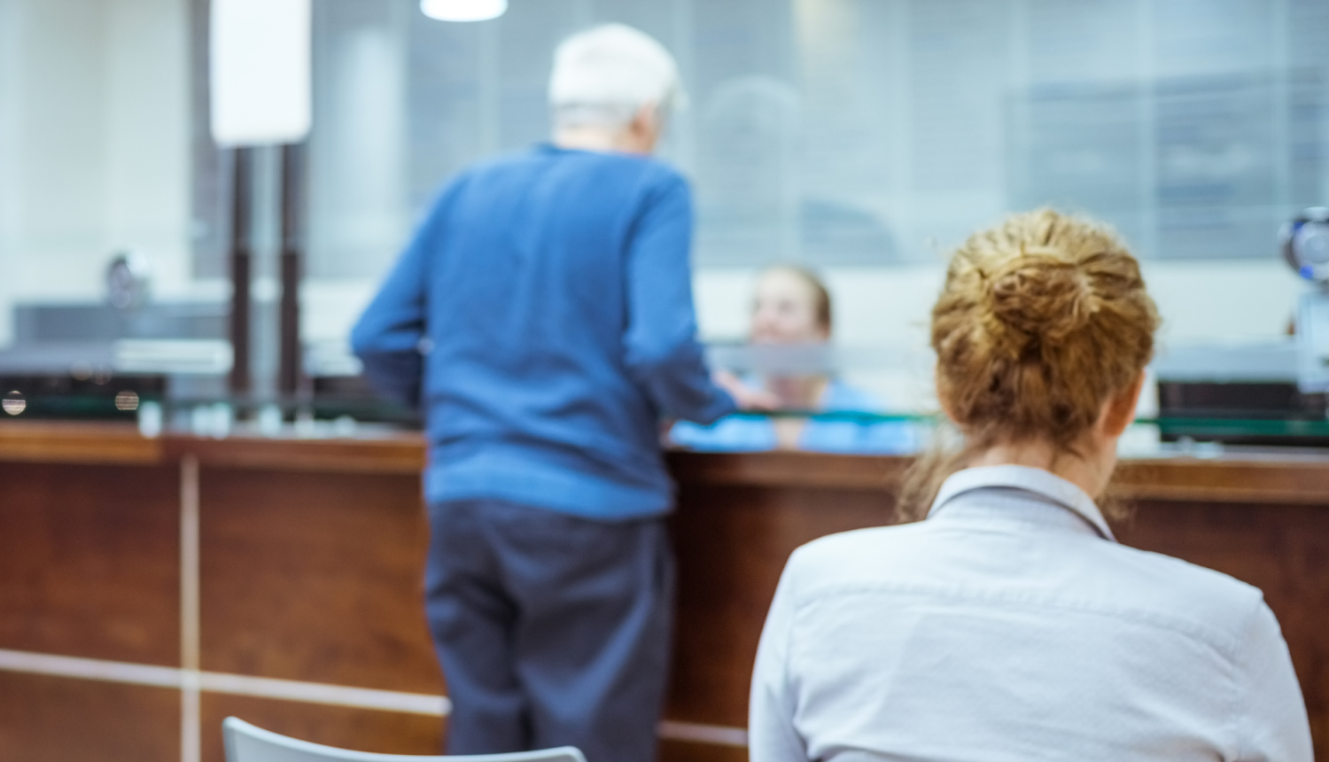 Klients pie kases vai klientu apkalpošanas lodziņa bankā, pastā vai valsts iestādē, fonā redzama darbiniece, bet priekšplānā – cita kliente no mugurpuses.