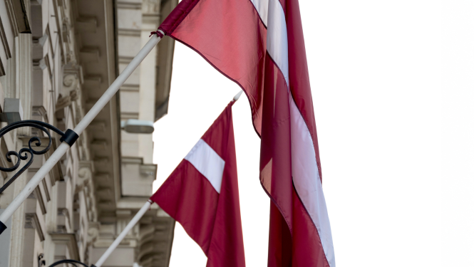 Divi Latvijas valsts karogi pie ēkas fasādes, kas plīvo vējā uz gaiša debesu fona.