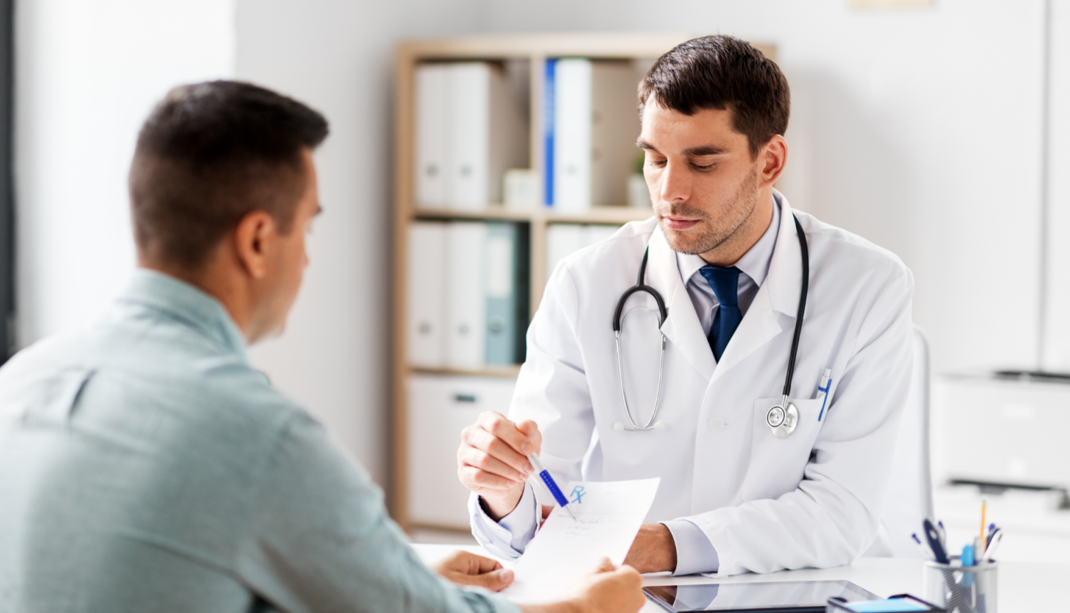 Ārsts vīrietis sēž pie galda pretī pacientam un ar pildspalvu norāda uz informāciju veidlapā