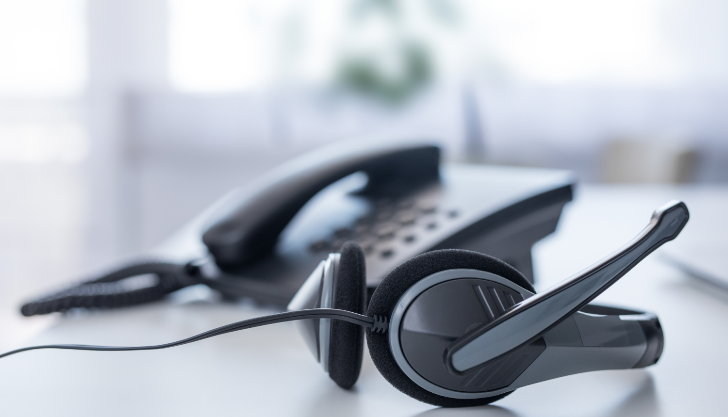 Biroja austiņas ar mikrofonu un stacionārais tālrunis uz balta galda.